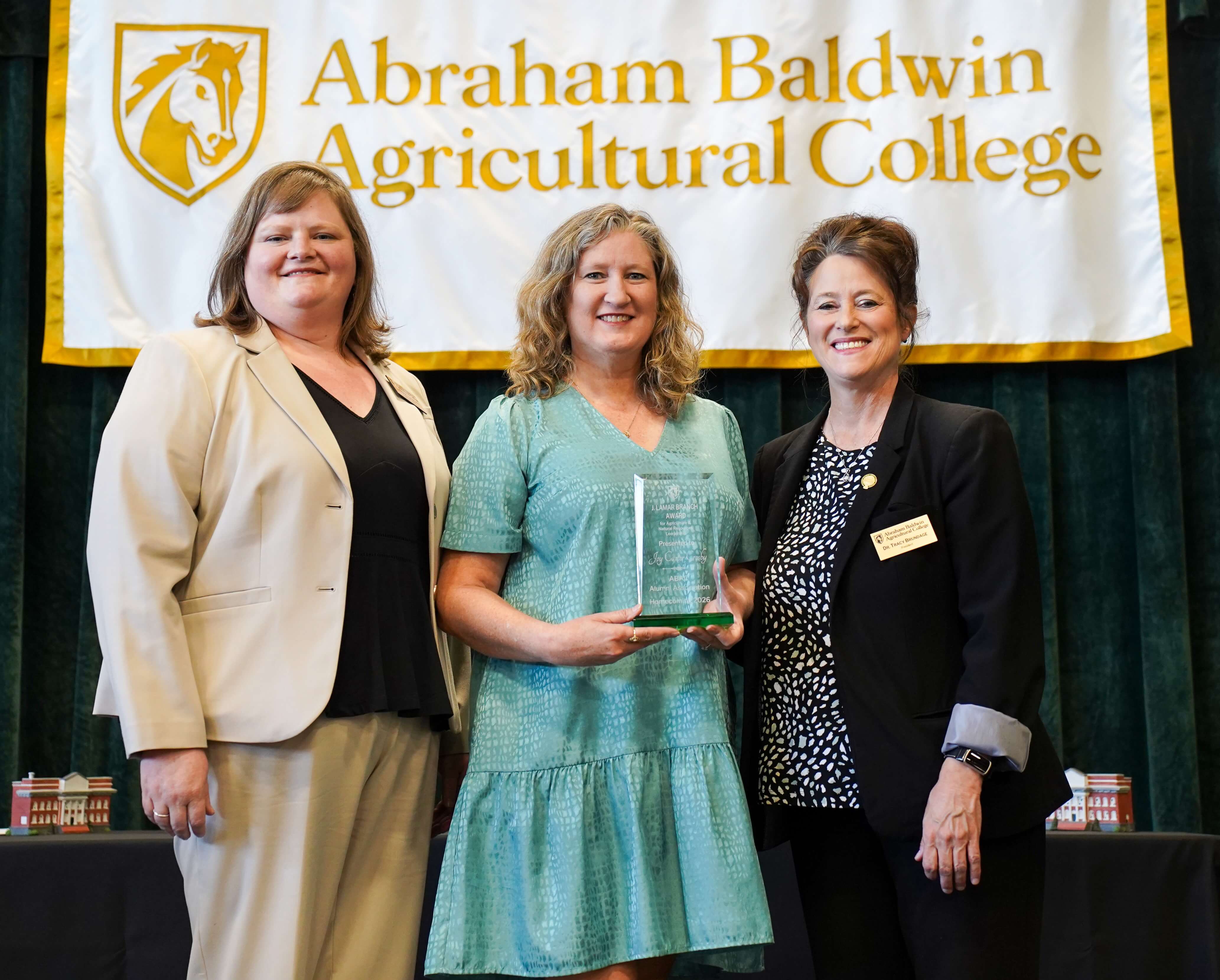 three people at an award presentation on stage with the winner holding her award.